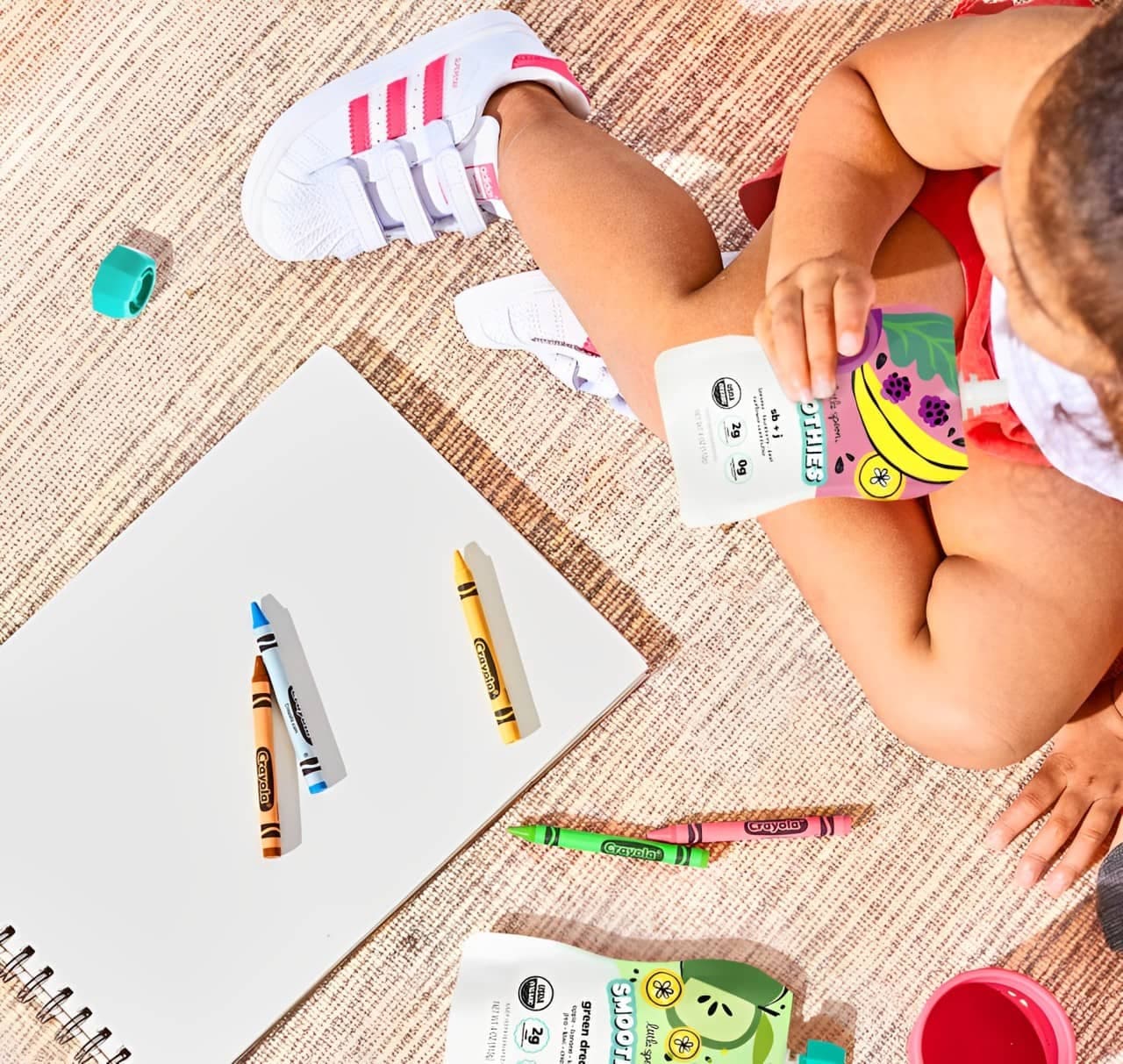 Baby sitting with crayons and a notebook while drinking a Little Spoon Smoothie pouch
